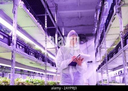 Portrait of female agricultural engineer writing on clipboard while standing between shelves in plant nursery greenhouse lit by blue light, copy space Stock Photo