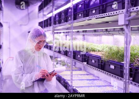 Waist up portrait of female agricultural engineer writing on clipboard while standing between shelves in plant nursery greenhouse lit by blue light, copy space Stock Photo