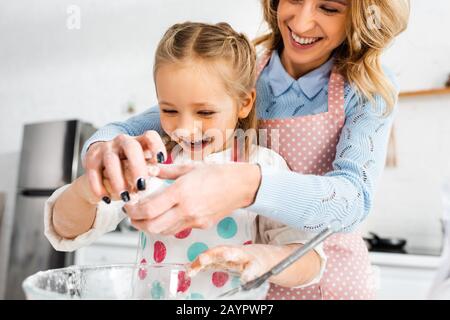 Selective focus of attractive mother and cute daughter laughing during adding  ingredients to dough in bowl Stock Photo