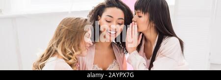 panoramic shot of smiling multicultural girlfriends whispering and gossiping on pajama party Stock Photo