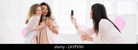 panoramic shot of cheerful girl taking photo of multicultural girlfriends with glasses of champagne on smartphone during pajama party Stock Photo