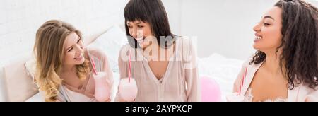 panoramic shot of multicultural girlfriends and laughing pregnant woman holding glasses with milkshakes on baby shower Stock Photo