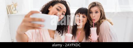 panoramic shot of happy multicultural girlfriends in bathrobes drinking milkshakes while taking selfie on smartphone during bachelorette party Stock Photo