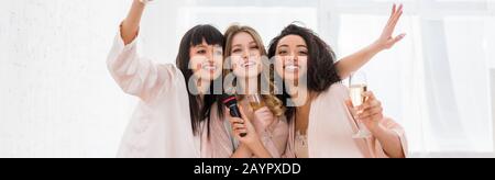 panoramic shot of attractive cheerful multicultural girls holding champagne and singing with microphone on bachelorette party Stock Photo