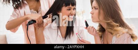 panoramic shot of smiling multicultural girlfriends doing hair styling and makeup with lipsticks on bachelorette party Stock Photo