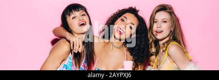 panoramic shot of happy fashionable multiethnic girls holding glasses with milkshakes on pink Stock Photo
