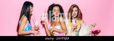 panoramic shot of cheerful stylish multiethnic girlfriends drinking milkshakes on pink Stock Photo