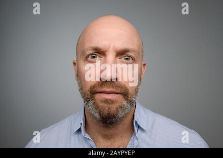 Keep calm and pretend nothing happened. Mature male with beard standing with no emotions over gray background, staring at camera without interest. Stock Photo