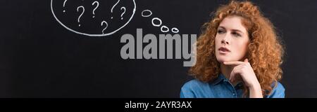 panoramic shot of beautiful thoughtful redhead girl with question marks in thought bubble on blackboard Stock Photo