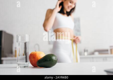 Selective focus of glass of water and fruits on table and smiling sportswoman measuring waist with tape in kitchen Stock Photo