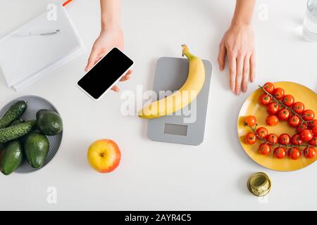 Top view of girl holding smartphone while weighing banana on kitchen table, calorie counting diet Stock Photo