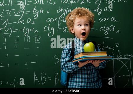 surprised kid holding books and apple near chalkboard with mathematical formulas Stock Photo