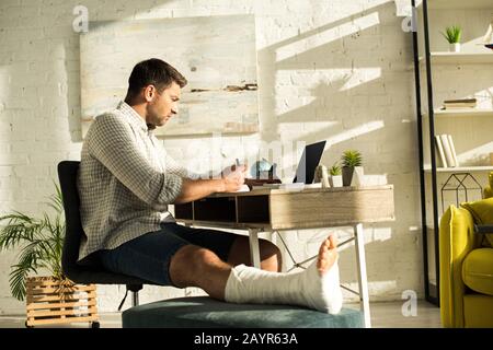 Side view of handsome freelancer with broken leg writing on notebook and using laptop at table Stock Photo