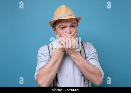 Senior mature, shocked, surprised man with hands covering mouth, eyes wide open in full disbelief, isolated on blue background. Stock Photo