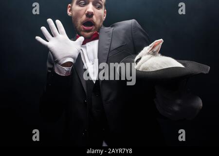 scared magician holding white rabbit in hat, isolated on black Stock Photo