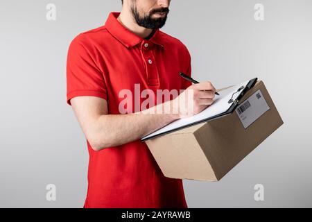 Cropped view of delivery man writing on clipboard and holding cardboard box isolated on grey Stock Photo