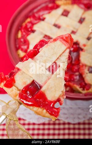 Piece of cherry pie being lifted out of a whole cherry pie, with a pie server with a ribbon on it.  Background is a red and white checkered tablecloth Stock Photo