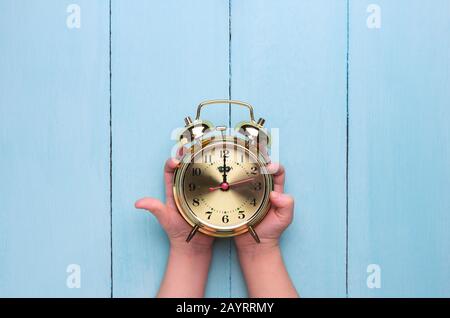 Golden alarm clock in children's hands on a light blue background. Time concept, top view, place for text. Stock Photo