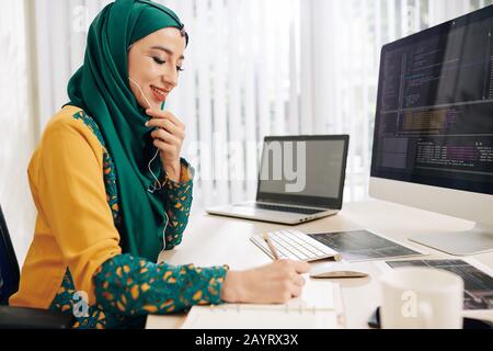 Happy young Asian freelance software developer talking on phone with client and taking notes in planner Stock Photo