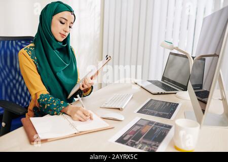 Female software engineer checking docuDFHJDHGFJHSDFKHKJFHKDSJHFKJHDFKJDHts with printed out programming code and taking notes in notebook Stock Photo