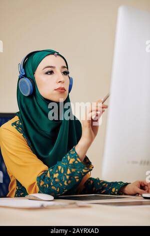 Serious female software developer in hedphones searching for mistakes in programming code on computer screen Stock Photo