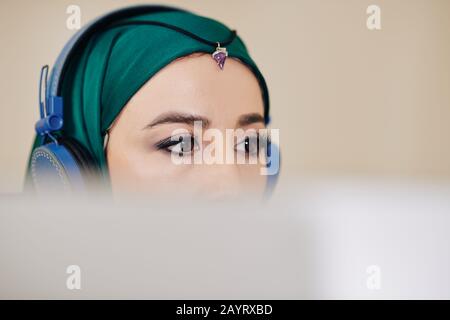 Young beautiful software engineer in hijab listening to music in headphones when programming or editing video on computer Stock Photo