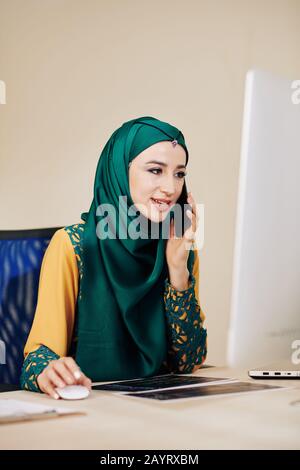 Portrait of young muslim software engineer talking on phone with client and checking details of project Stock Photo