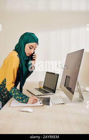 Musilim software engineer talking on phone with project manager and writing down his ideas in notepad Stock Photo