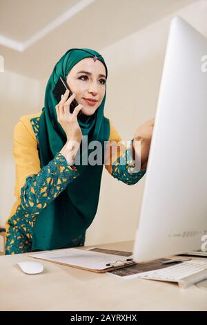 Smiling pretty female engineer talking on phone when discussing strings of code on somputer screen Stock Photo
