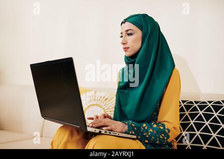 Muslim female freelance programmer sitting on sofa at home and working on laptop Stock Photo