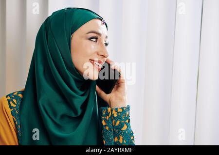 Beautiful smiling young muslim woman in deep green hijab looking through blinds on window and talking on phone Stock Photo