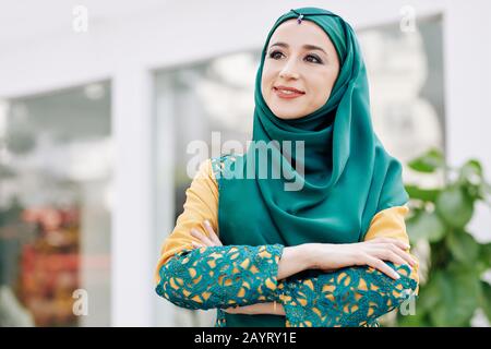 Portrait of young positive pensive woman in hijab standing outdoors with arms folded Stock Photo