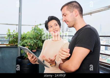 Creative young Vietnamese woman with digital tablet showing presentation of her creative idea to coworker Stock Photo