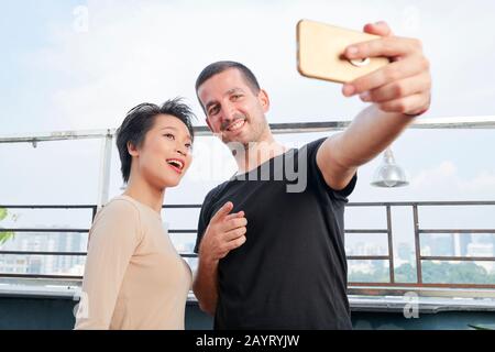Happy multi-ethnic friends or colleagues standing on rooftop and talking selfie for social media Stock Photo