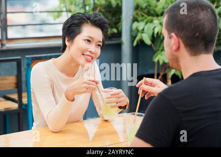 Beautiful young Vietnamese woman enjoying citrus cocktail when looking at her friend telling interesting story Stock Photo
