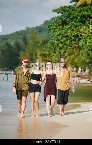 Two smiling couples in sunglasses hugging when walking on tropical beach together Stock Photo