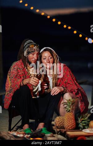 Couple covering with blanket and drinking bottled beer when spending night at fireplace on the beach Stock Photo