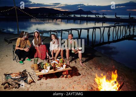 Group of friends resting on beach at night, playing ukulele and esting food by fireplace Stock Photo