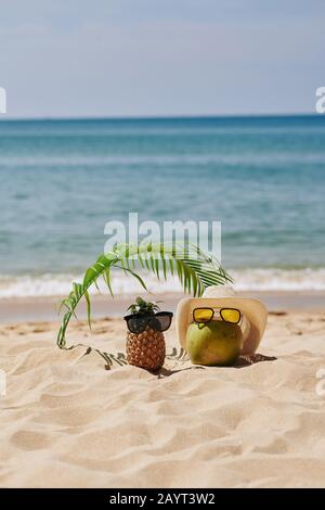 Coconut and pineapple in sunglasses resting under palm leaf on the beach Stock Photo