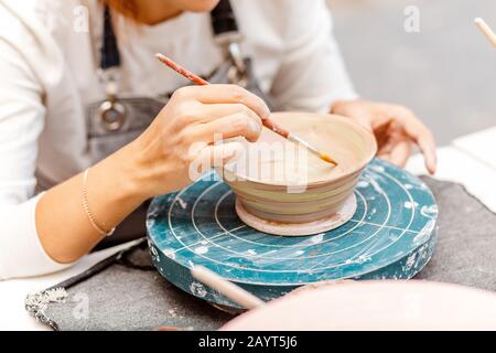 A woman artist in working apron paints a clay pottery among brushes and other tools in the modern workshop Stock Photo