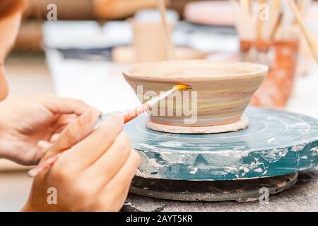 A woman artist in working apron paints a clay pottery among brushes and other tools in the modern workshop Stock Photo