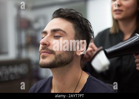Stylish man sitting barber shop Hairstylist Hairdresser Woman blow dry his hair Portrait handsome happy young bearded caucasian guy getting trendy hai Stock Photo