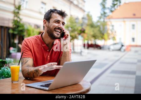 Smliling man talking on a phone while sitting in front of a laptop in cafe Stock Photo