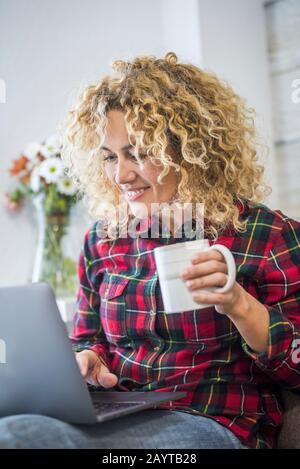 Happy cheerful pretty young woman with blonde curly hair work and smile with laptop computer sit down on the sofa at home - concept of modern people a Stock Photo