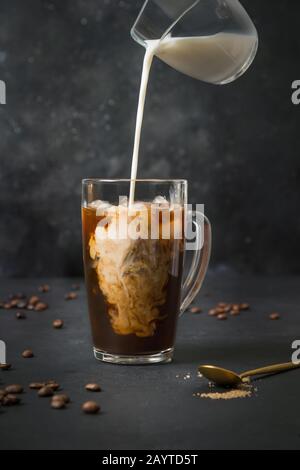 Iced latte coffee in cup glass with pouring milk on black. Close up. Stock Photo