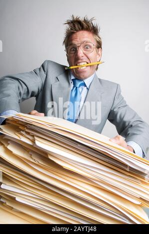 Stressed businessman with pencil in his mouth trying to lift a massive pile of paperwork on his desk Stock Photo
