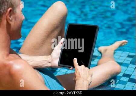 Over the shoulder view of man relaxing with tablet computer on the edge of bright blue tiled swimming pool Stock Photo