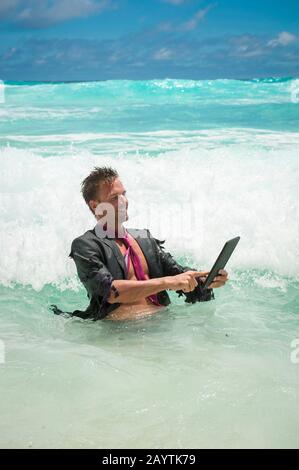 Cheerful castaway businessman using a tablet computer in tropical waves Stock Photo