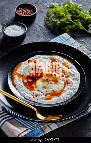 close-up of cilbir, poached eggs in yogurt sauce topped with spicy  butter served on black plates with golden cutlery, turkish cuisine Stock Photo
