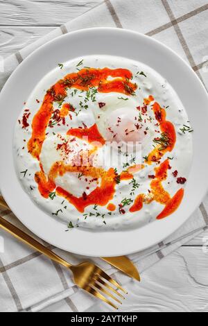 Turkish eggs with greek yogurt and aleppo pepper butter sauce, cilbir, served on white plates with golden cutlery on a white wooden table,  vertical v Stock Photo